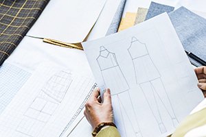 Designer holding fashion sketches of a sleeveless dress set surrounded by fabric swatches and patterns.