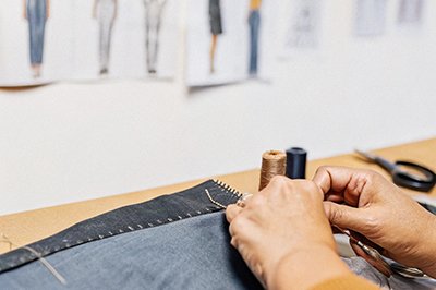 Close-up of a dressmaker hand stitching denim fabric with thread and needles, with sketches in background.