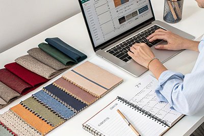 Designer working on laptop at desk with fabric swatches, planner, and color samples neatly arranged.