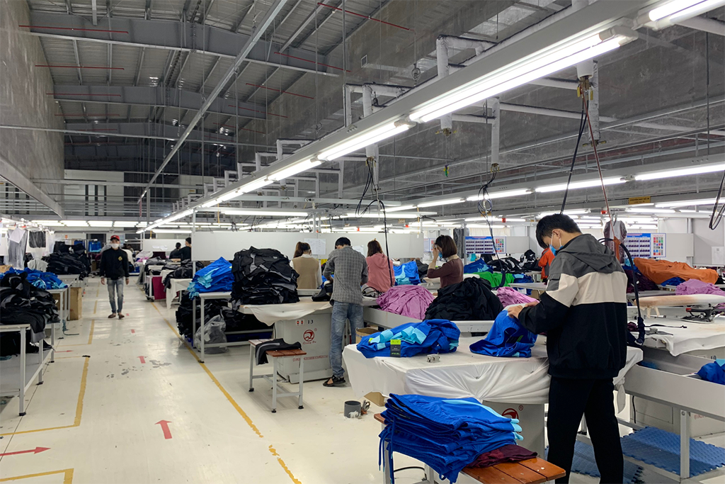 Workers in a brightly lit garment factory assembling and inspecting clothing on large workstations with fabric piles.
