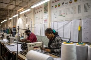 Workers sewing garments in a factory with production charts and equipment in the background.